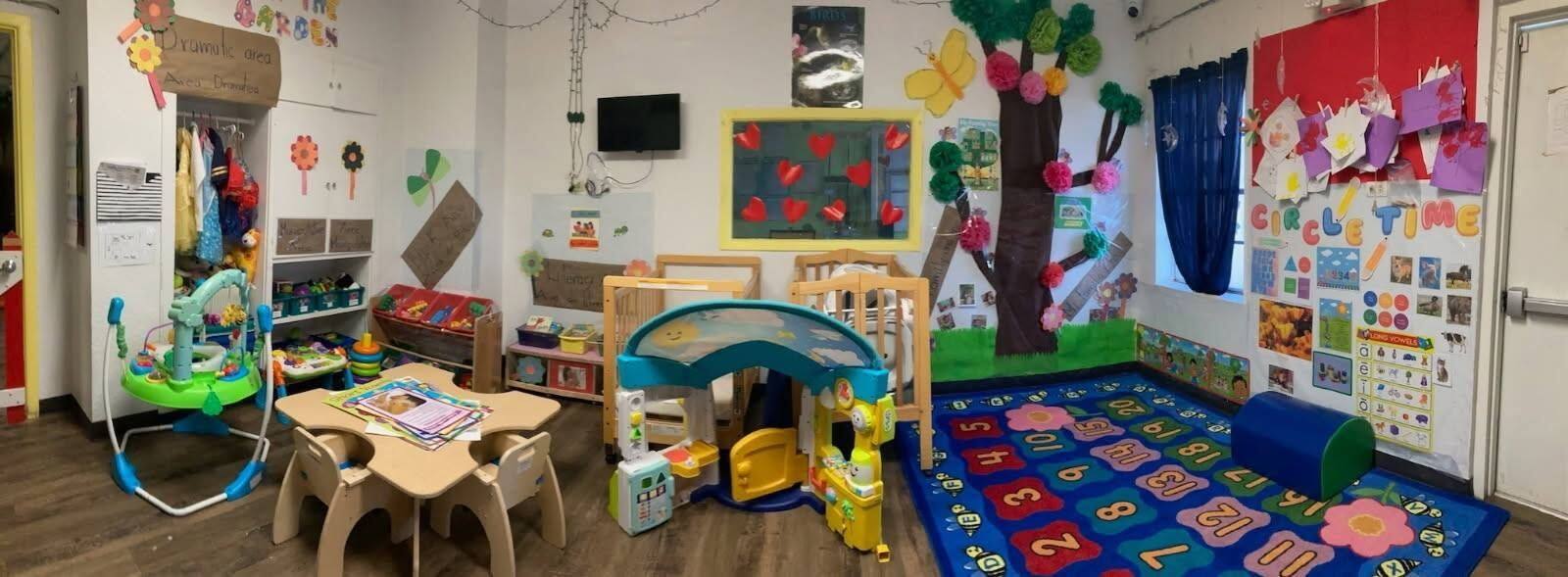 A bright infant classroom with cribs, toys, and a circle-time rug